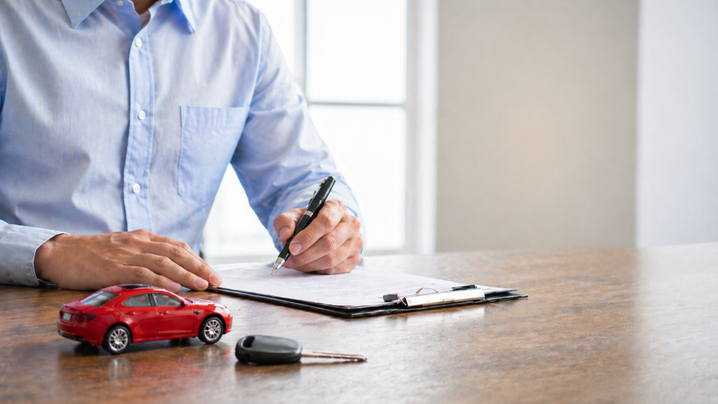 Person signing car insurance paperwork with car keys on a desk