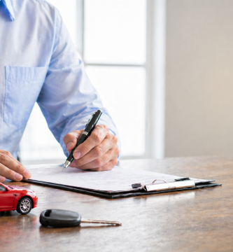 Person signing car insurance paperwork with car keys on a desk