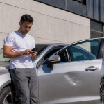 Driver checking a car insurance claim on a phone next to his parked car