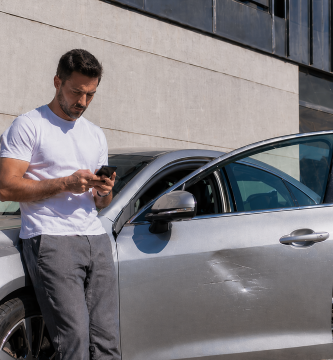 Driver checking a car insurance claim on a phone next to his parked car